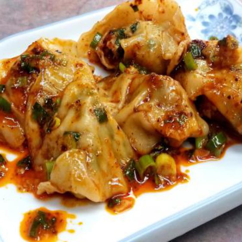 Taiwanese dumplings coated in a spicy orange sauce with green onions on a white plate.