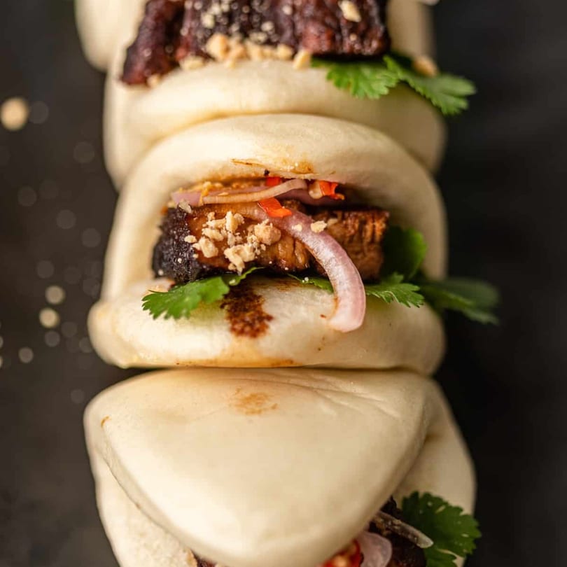 Three Taiwanese gua bao buns stacked, filled with pork belly, cilantro, red onion, and peanuts.